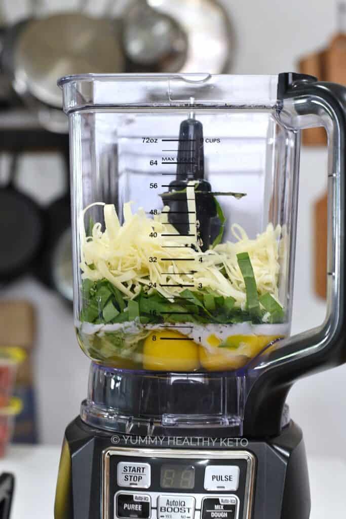 Eggs, milk, spinach and shredded cheese in a blender with pans and wooden cutting boards hanging on the wall in the background 