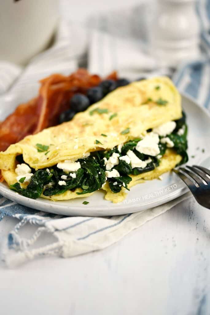 A close up image of a Spinach and Feta Omelette on a small plate with bacon slices and fresh blueberries.